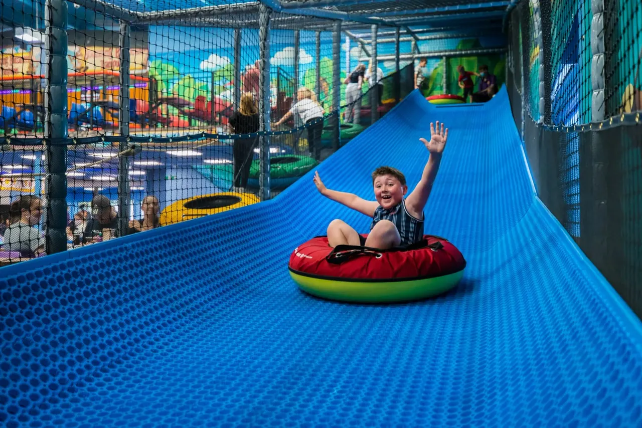 Child sledging down a blue indoor slide on a red and green inflatable tube with arms raised.
