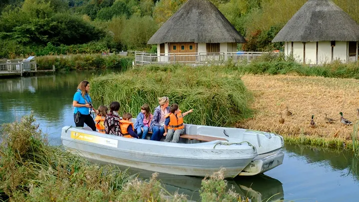 Adult and children in a boat on a river with thatched-roof visitor buildings and nature reserve in background.