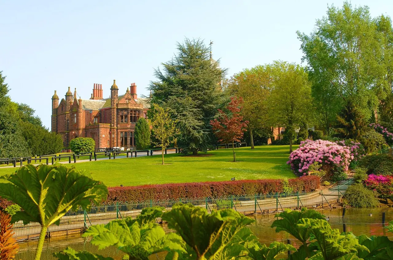 Victorian brick mansion with turrets set in manicured gardens with flowering shrubs, trees and lawn beside a fenced waterway.