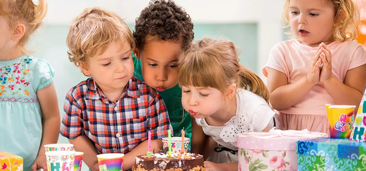 Group of young children gathered around a birthday cake with lit candles at a party celebration.