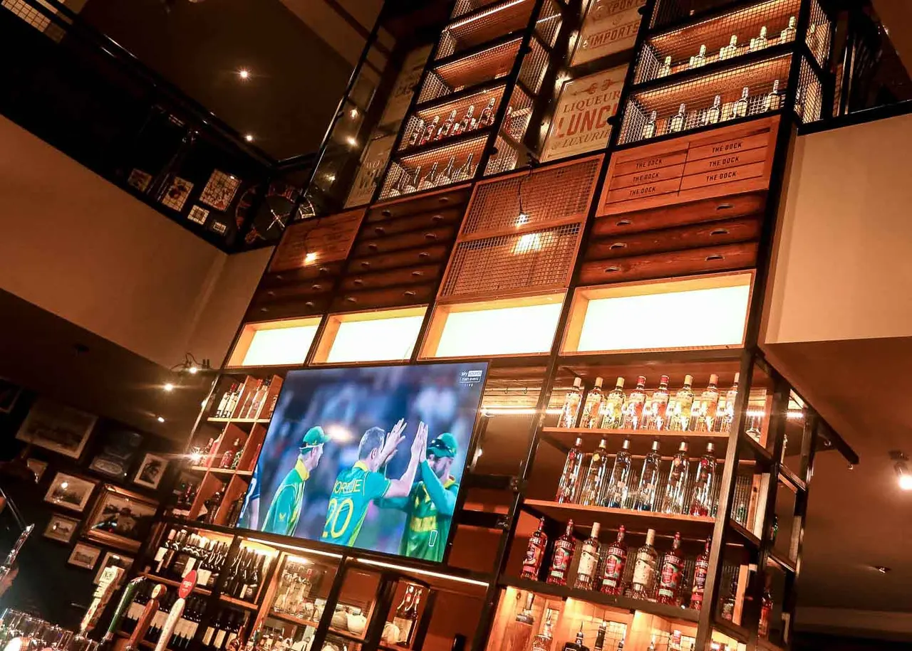 Interior of a sports bar with illuminated shelving displaying bottles, a TV screen showing cricket players, and warm wood panelling throughout.