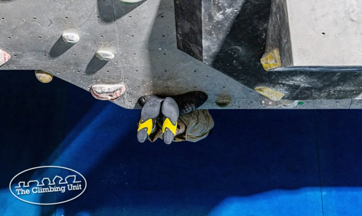 Overhead view of a climber's feet in yellow and black climbing shoes on an indoor climbing wall.