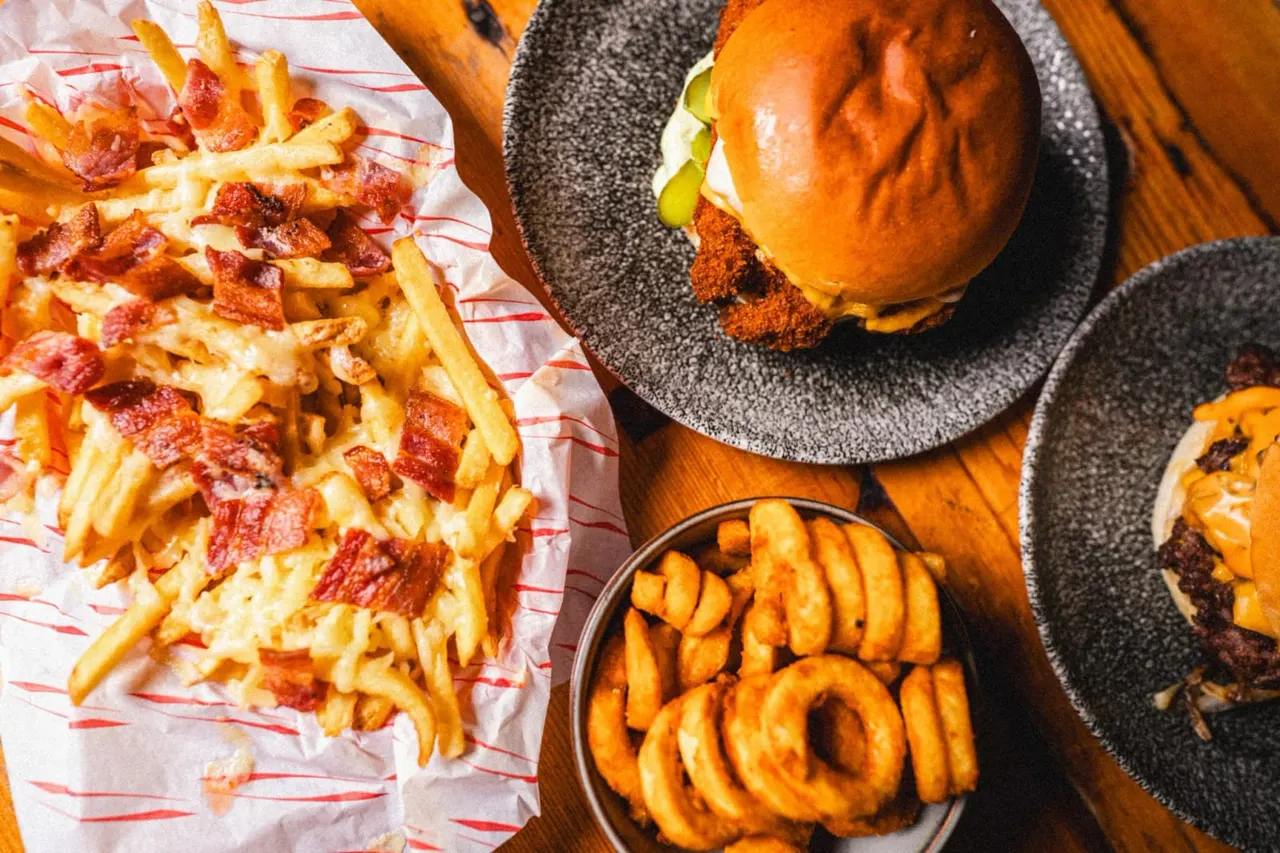 Bacon-topped chips, spiced chicken burger with pickles on a dark plate, and golden onion rings in a bowl on a wooden table.