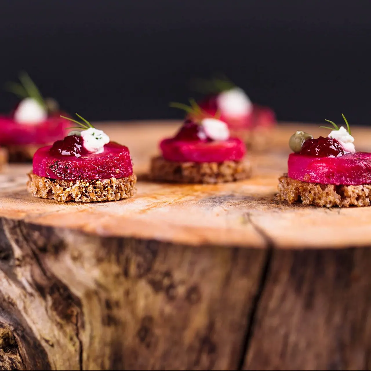 Small pink and red layered desserts with biscuit bases topped with berries and cream on a wooden board.
