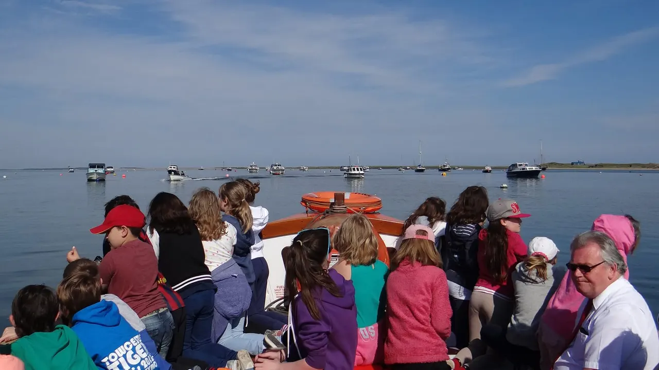 Passengers crowded on an orange-and-white boat touring a calm bay dotted with moored vessels under blue sky.