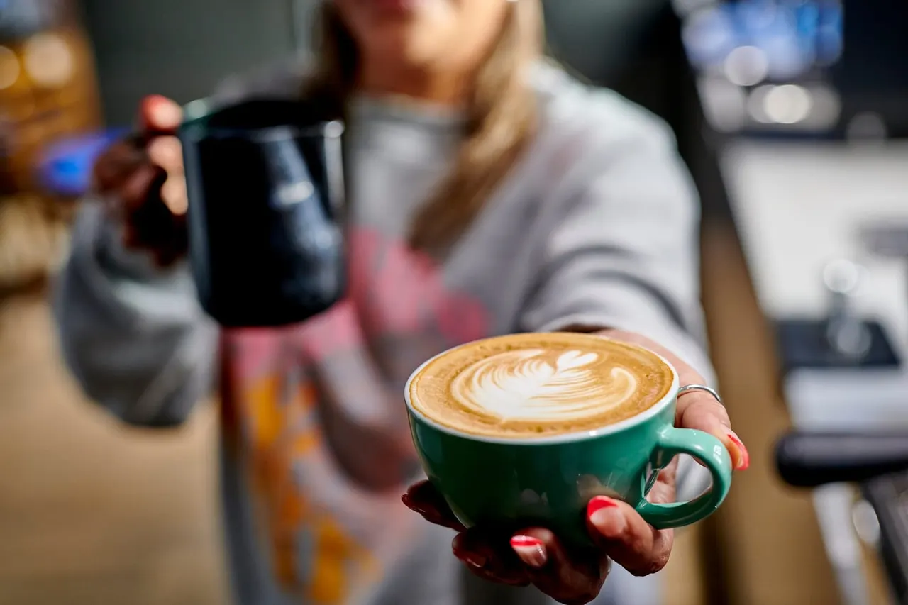 Person holding a green cup of latte with latte art, with another drink in the background.