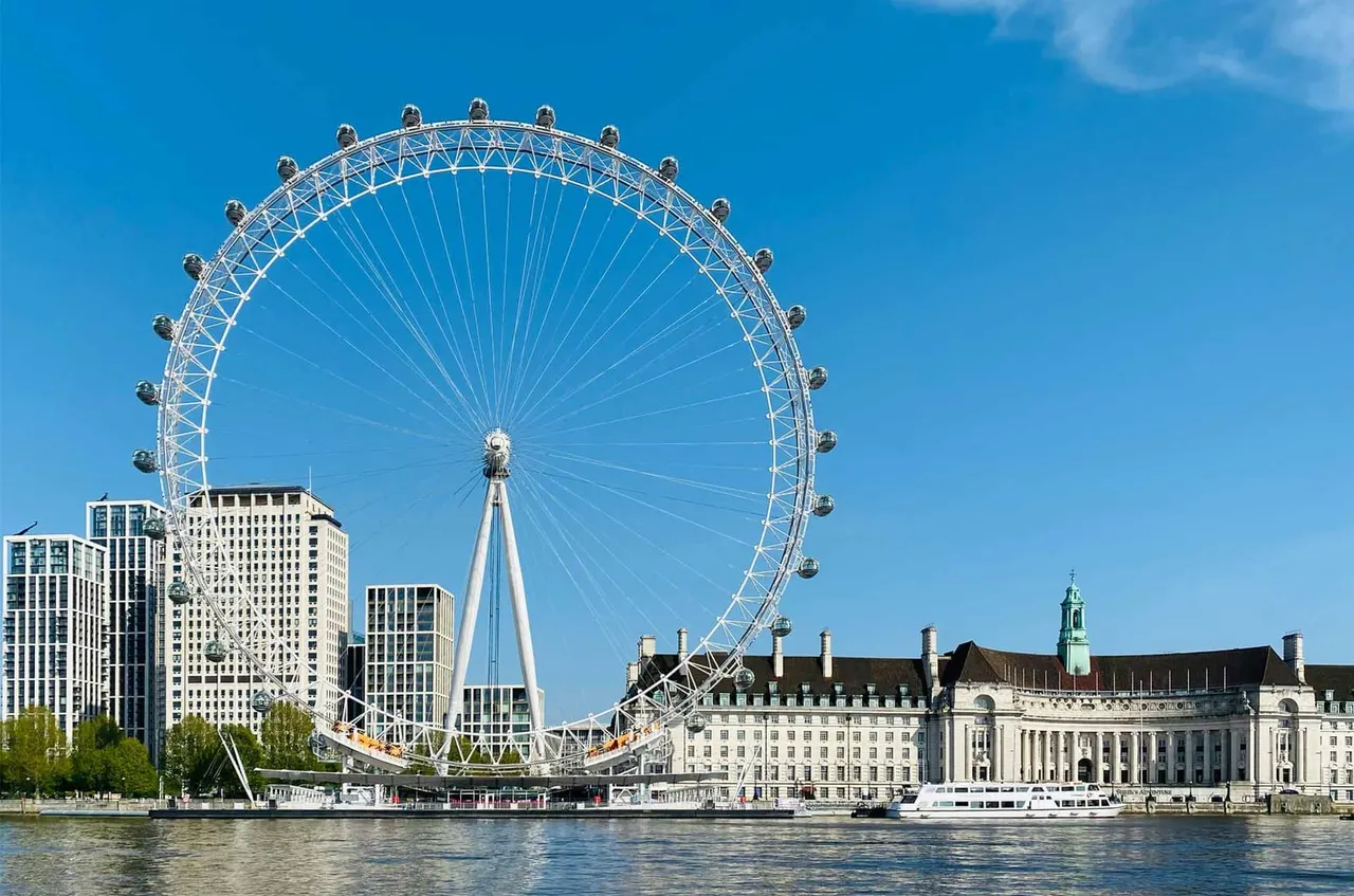 A large observation wheel on the South Bank of the River Thames with historic buildings and modern high-rises alongside, viewed across the water on a clear day.
