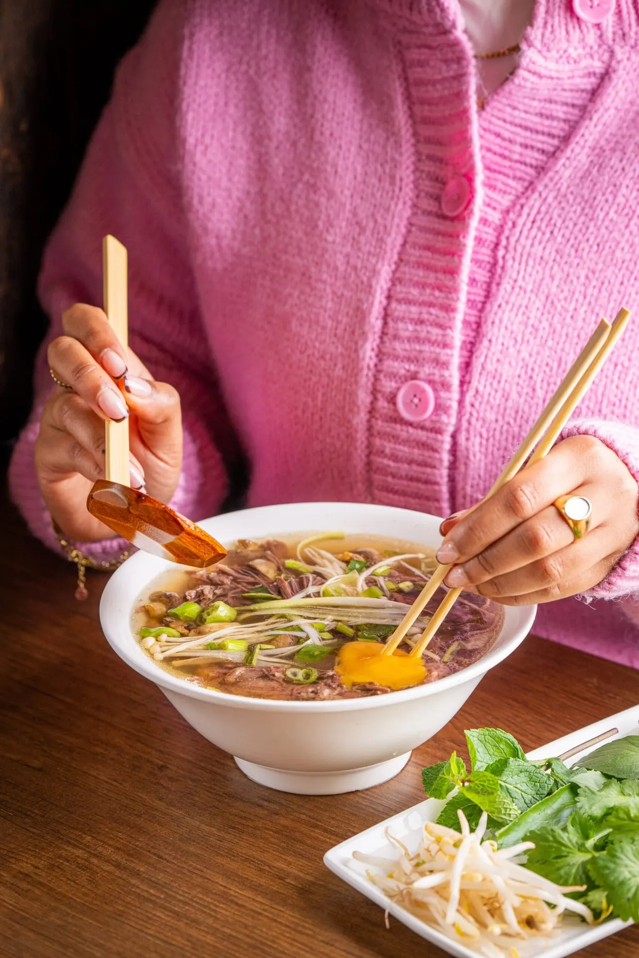 Person in pink cardigan eating noodle soup with chopsticks and spoon at a table.