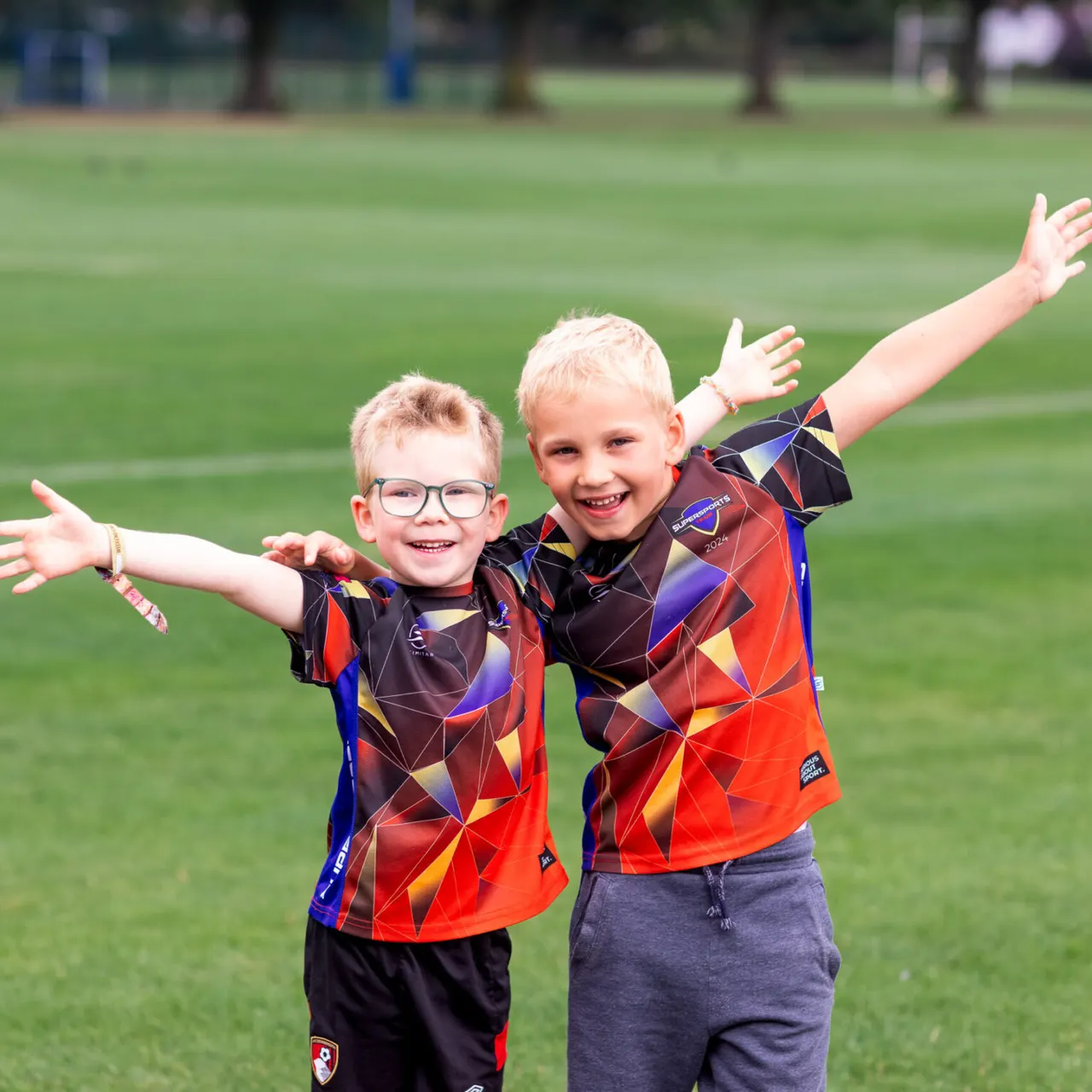 Two young boys in red and black sports jerseys with arms outstretched, smiling on a grass pitch.