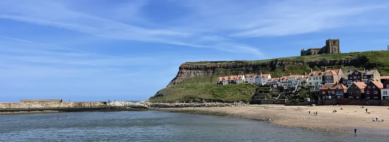 Seaside village nestled beneath a cliff with a castle ruin on top, sandy beach and visitors in foreground.