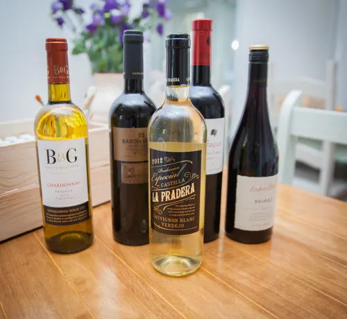 Five wine bottles displayed on wooden surface with purple flowers blurred in background.