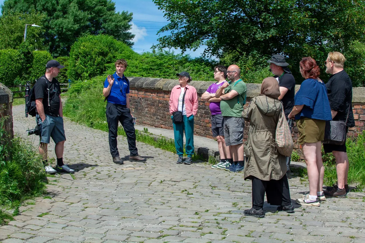A tour guide in a blue shirt addresses a group of visitors gathered on a paved pathway beside a brick wall beneath trees.