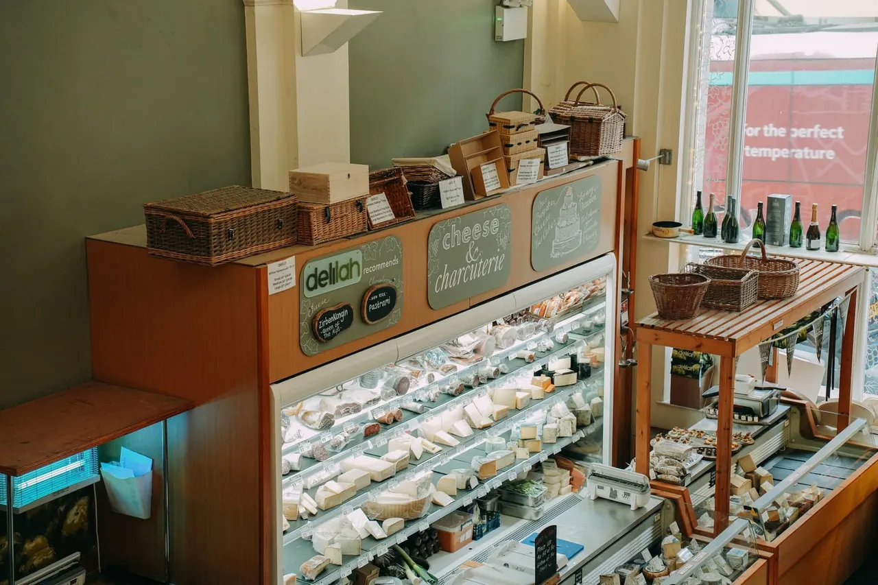 Interior of a delicatessen counter displaying various cheeses and charcuterie in refrigerated cases, with wicker baskets and wine bottles.