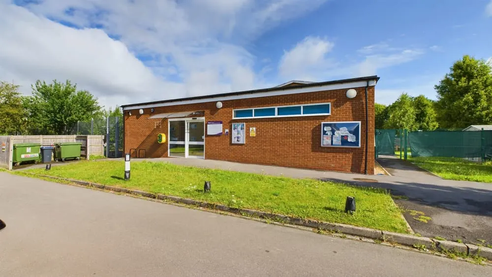 Modern brick building with glass entrance doors and blue windows, set back from car park with green lawn and trees.
