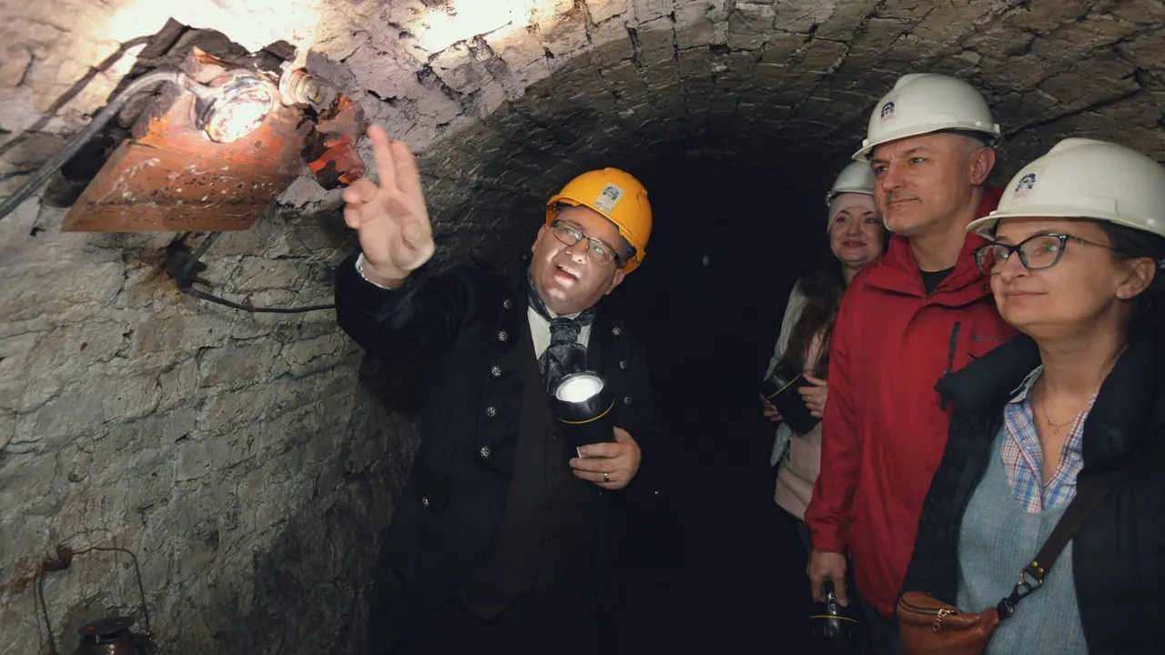 A guide in a yellow hard hat points out mining equipment to visitors wearing white hard hats inside a stone tunnel.