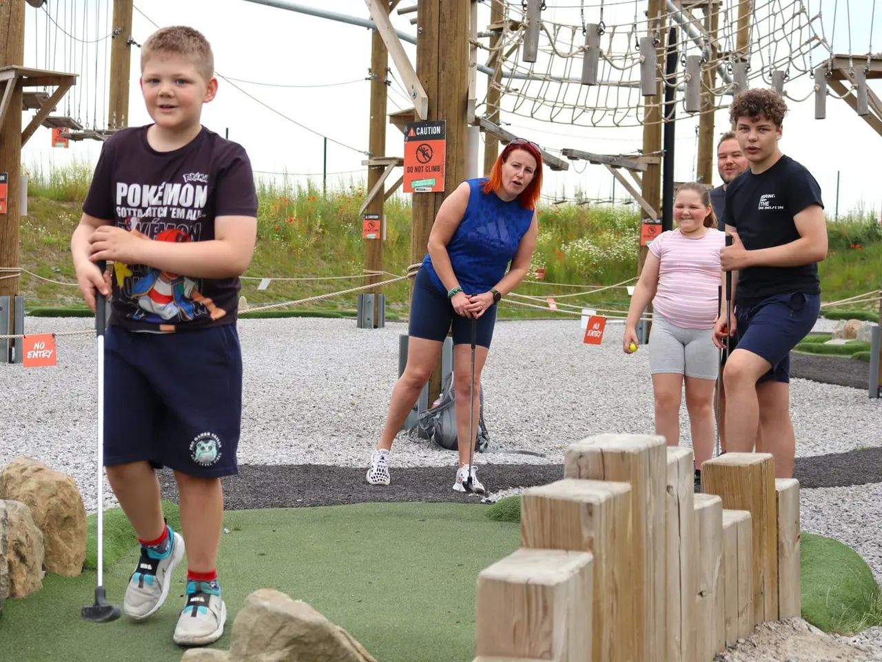 Children and an adult playing mini golf on an outdoor course with wooden obstacles and rope structures.