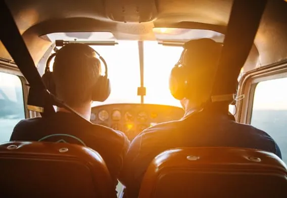 Two pilots in headsets sit in cockpit of small aircraft at sunrise or sunset.