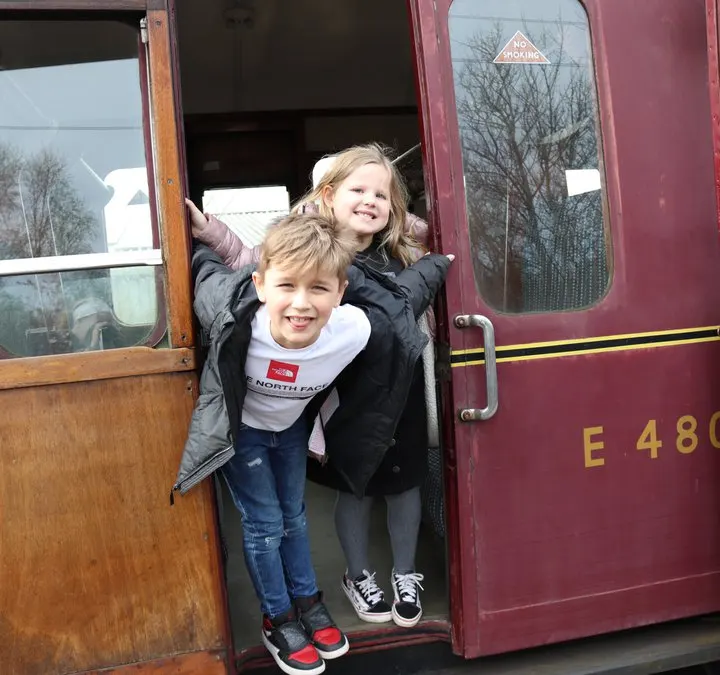 Two young children smiling in the doorway of a burgundy heritage railway carriage.
