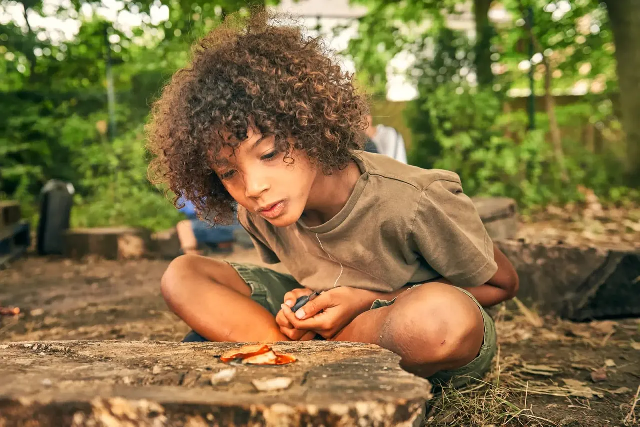 Child crouching on ground examining leaves and natural objects in a woodland garden setting.