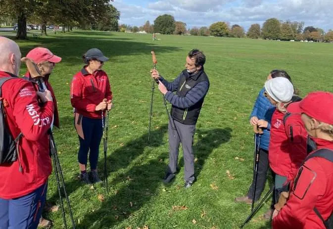 A group of people in red jackets and caps gather on a grassy park with Nordic walking poles for instruction.