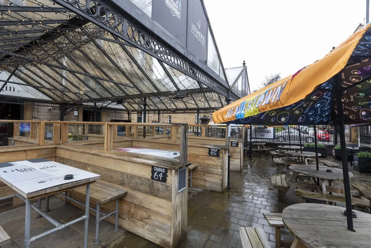 Outdoor covered dining area with wooden booth seating, picnic tables and colourful umbrellas under a Victorian iron and glass canopy.