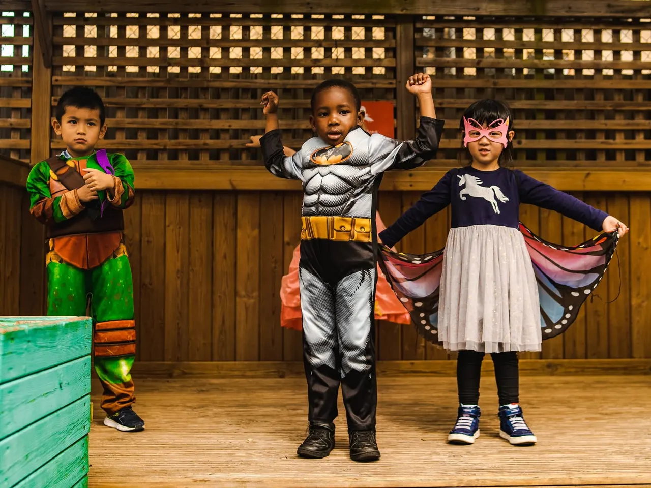 Three young children in superhero and fantasy costumes posing together in a wooden-walled indoor space.
