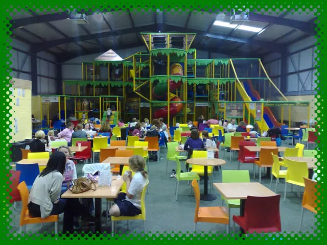 Children and adults seated at colourful tables in an indoor play centre with a large yellow and green soft play structure behind them.