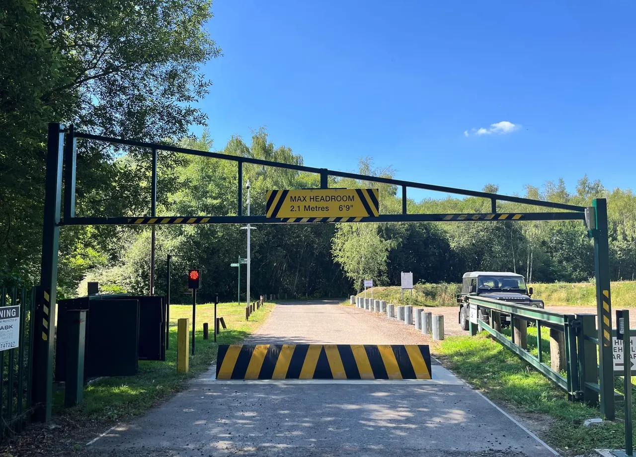 A vehicle approaching an entrance barrier with a maximum headroom warning sign, surrounded by woodland and green fields.