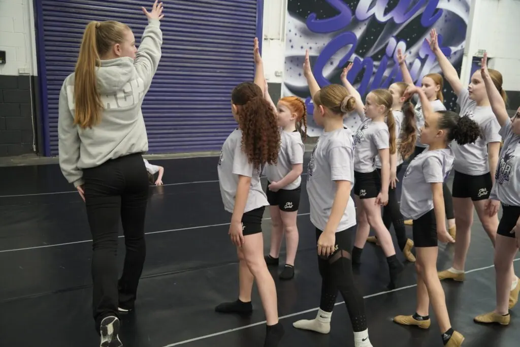 Instructor leading a group of young dancers in matching grey t-shirts during a dance class in a studio with purple signage.