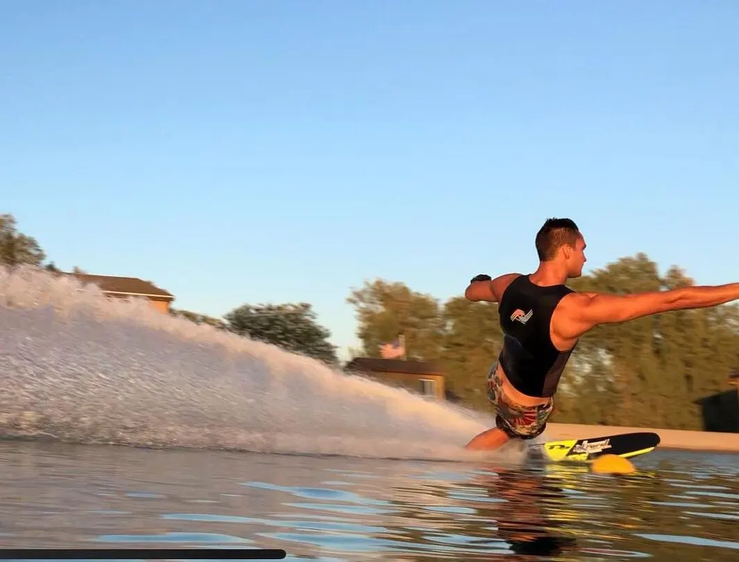 A man wakeboarding behind a boat, arms outstretched, towing a yellow wakeboard across water.