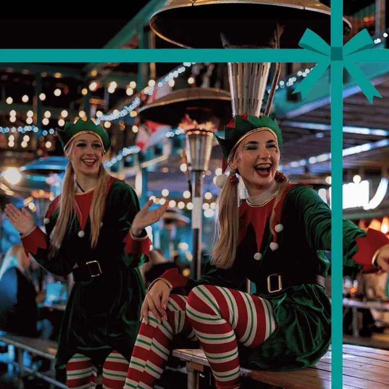 Two people in festive elf costumes smiling at a Christmas-themed venue with twinkling lights and decorations overhead.