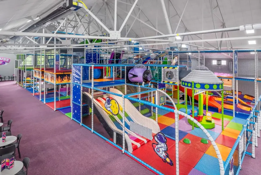 Colourful indoor soft play area with multiple levels, slides, climbing structures and padded equipment under white industrial ceiling.