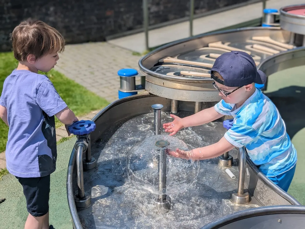 Two young children playing at an outdoor water table with pumps and flowing water on a sunny day.