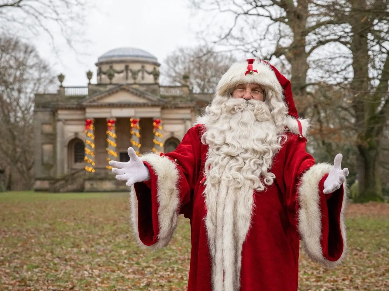 Person in Father Christmas costume with arms outstretched in front of historic stone chapel decorated with garland.