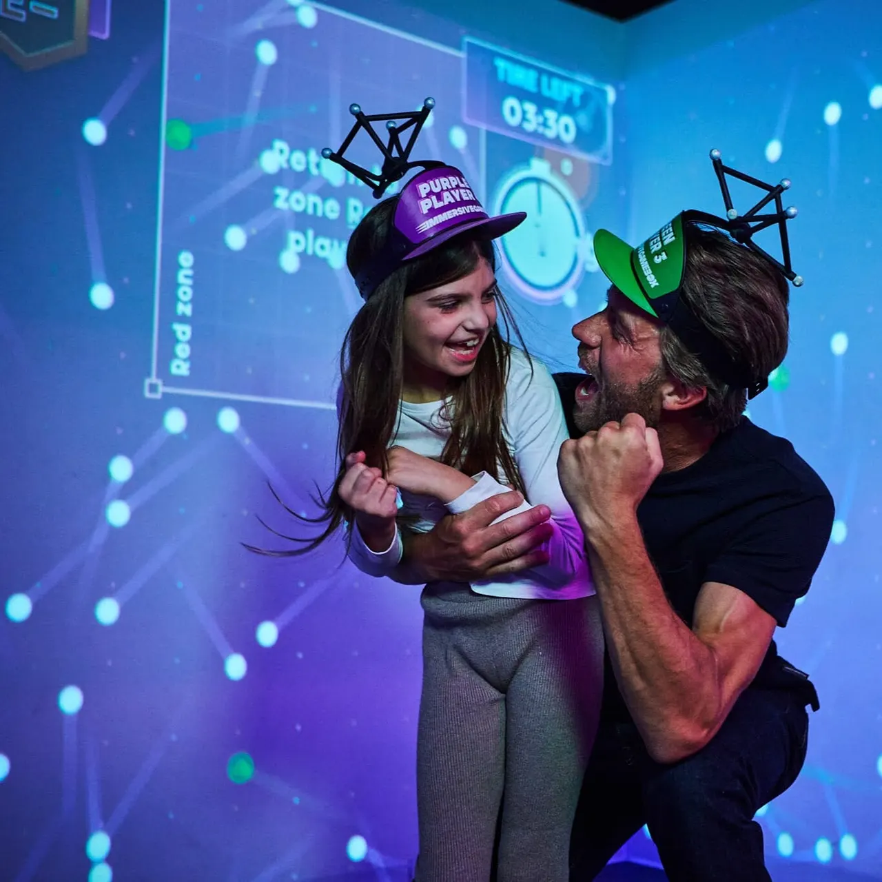 An adult and child wearing geometric headsets play together in a neon-lit gaming environment with digital displays.