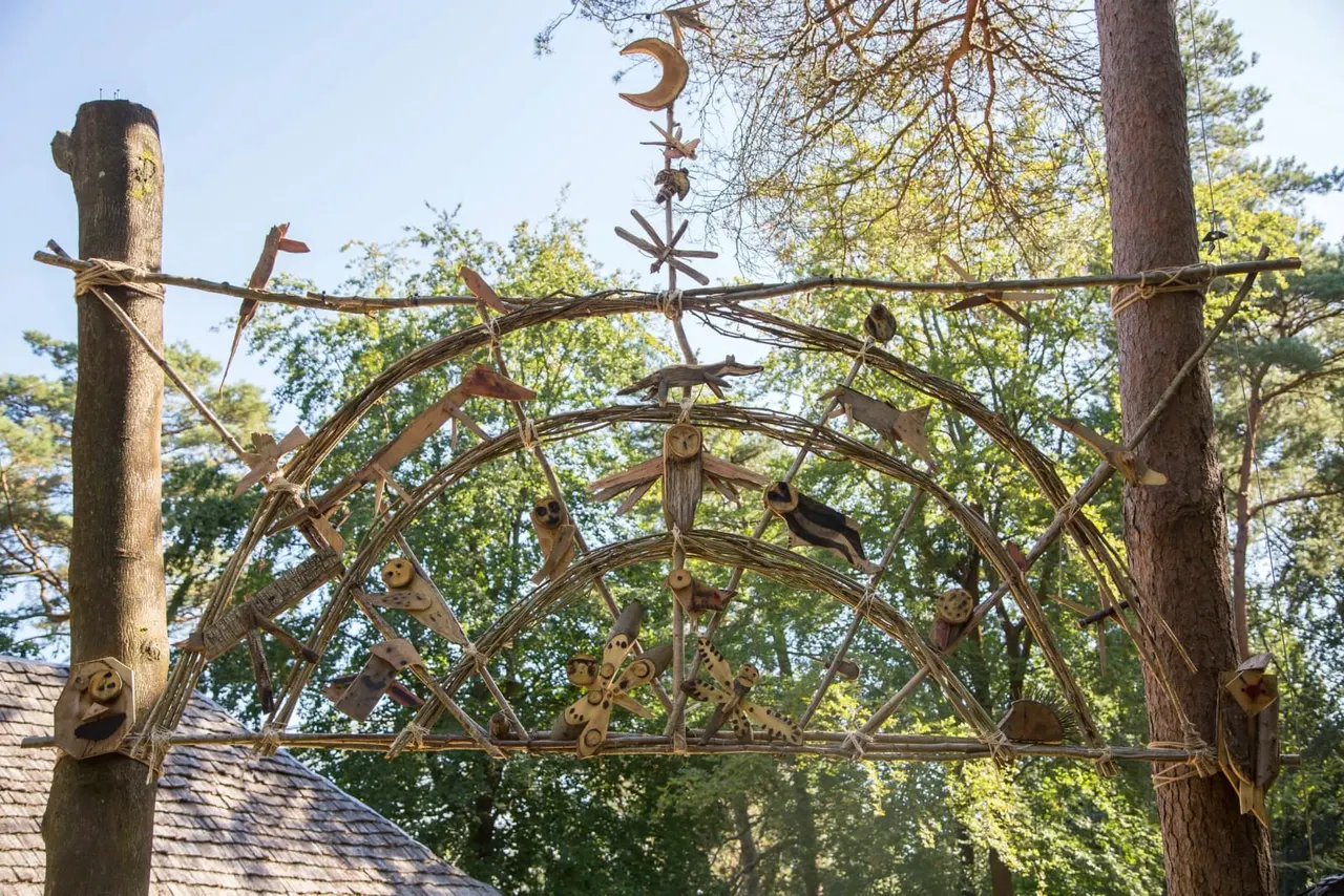 Decorative metal archway with woodland animal sculptures suspended between two tall wooden posts, surrounded by trees.