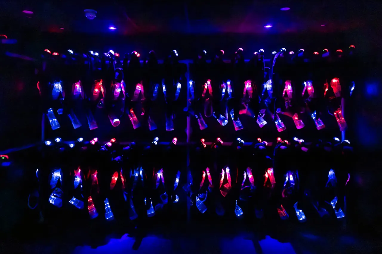 Large crowd of people wearing illuminated laser tag vests in red, blue and pink, holding laser guns in a darkened arena.