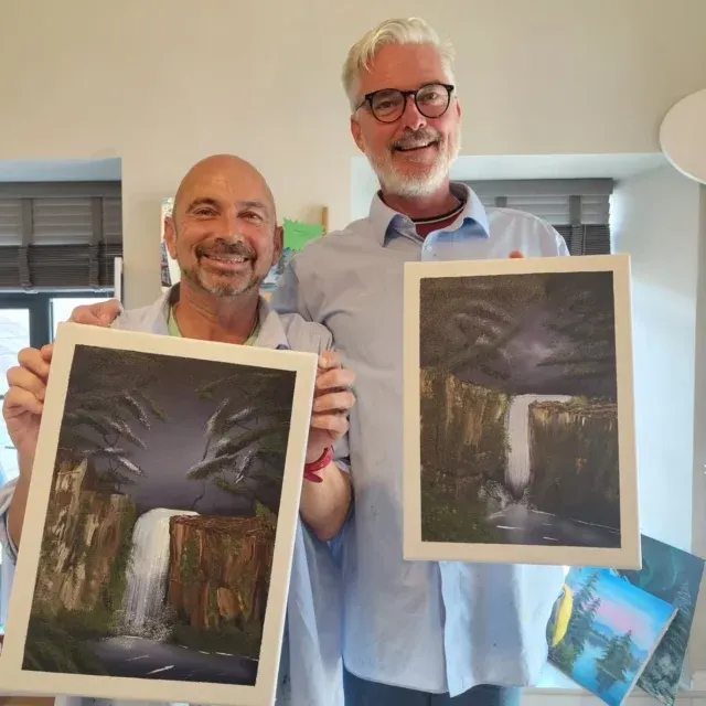 Two men holding framed paintings of waterfalls in a studio interior.