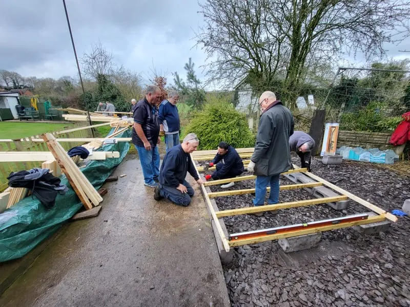 A group of volunteers working together to construct wooden raised garden beds filled with soil at an outdoor railway venue.