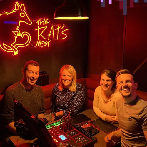 Four adults sit together at a table with gaming equipment, illuminated by a neon rat sign overhead.