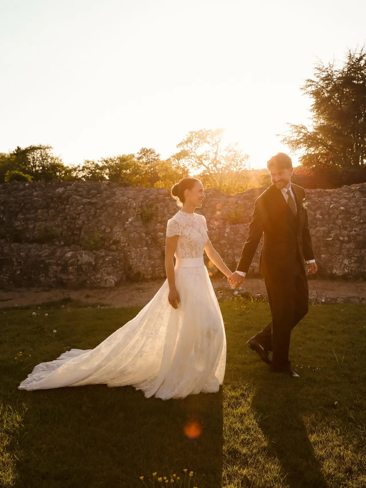 Bride and groom holding hands walking through grounds at sunset, with stone ruins and trees behind them.