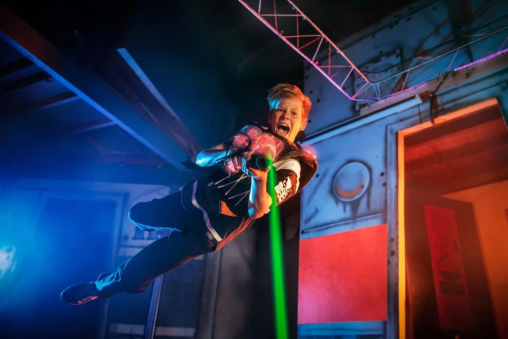 Player in neon combat gear firing laser gun during indoor laser tag game with colourful lighting.