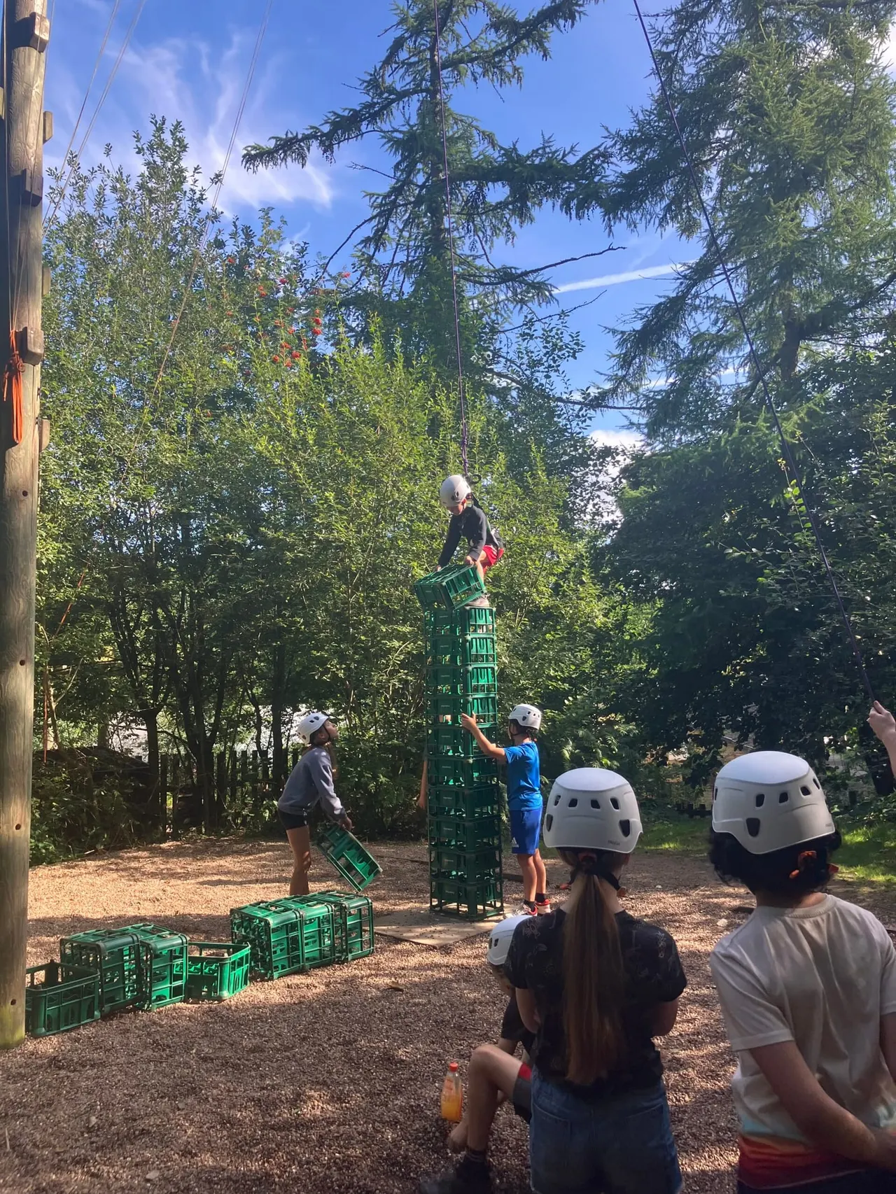 Children in safety helmets climbing a tall green tower structure made of crates in a woodland setting.