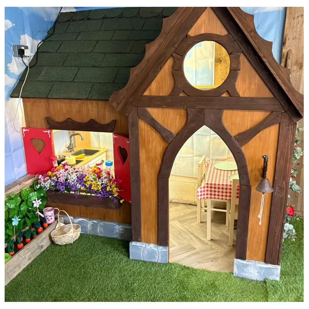 Decorated wooden playhouse with green roof, circular and arched windows, red door panel with sink, flower box, and interior furniture visible through open doorway.