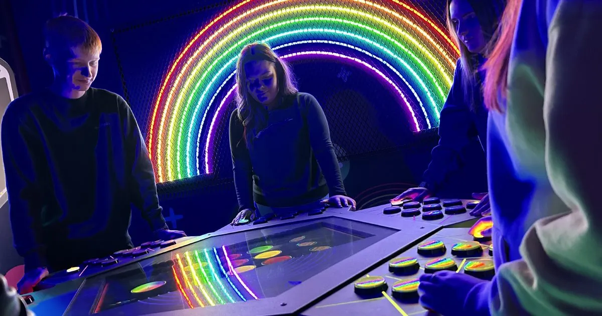 Young people playing an interactive glowing game table lit by a rainbow neon arch installation.
