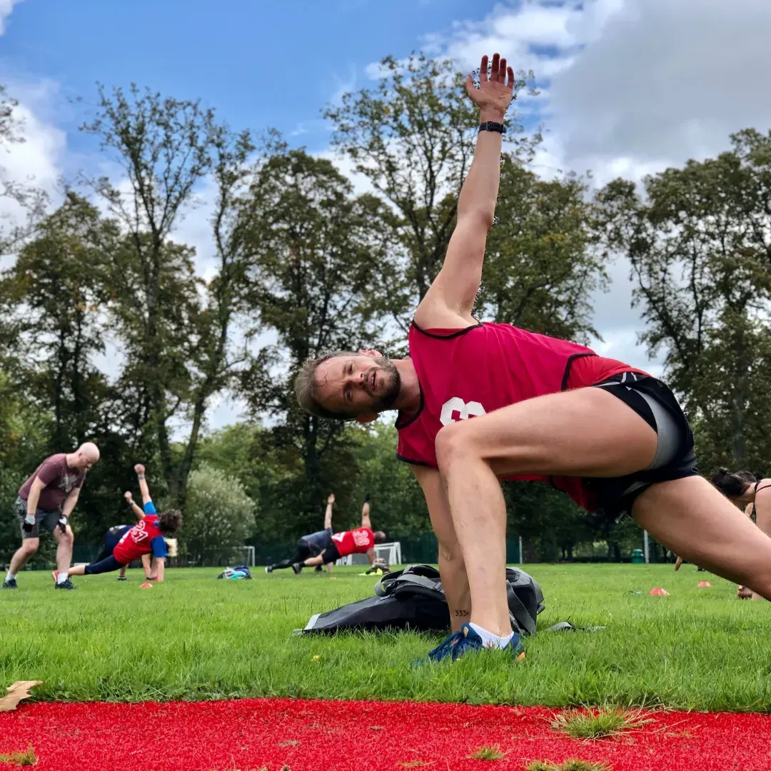 A person in a red shirt performs a side plank exercise on grass whilst others do fitness activities in the background at an outdoor park.