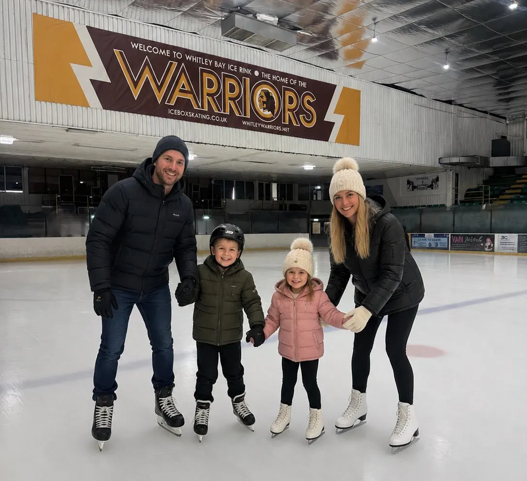 A family of two adults and two young children ice skating together at an indoor ice rink.