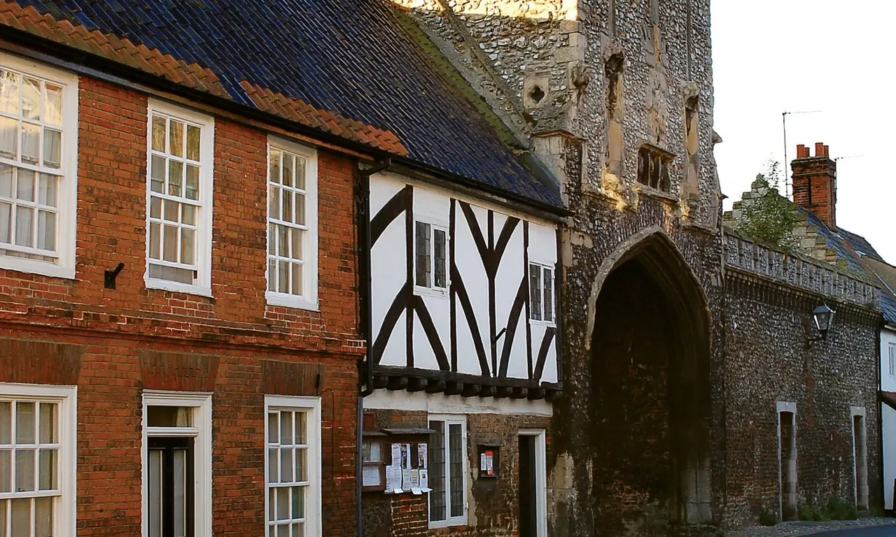 Historic timber-framed and brick buildings adjoining a stone church with Gothic arch gateway in a village street setting.