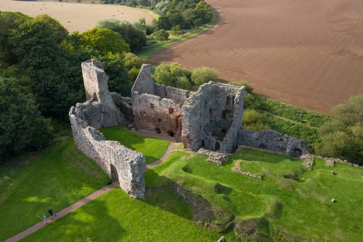 Aerial view of ruined medieval stone castle with thick walls and towers set on green grounds, surrounded by woodland and farmland.