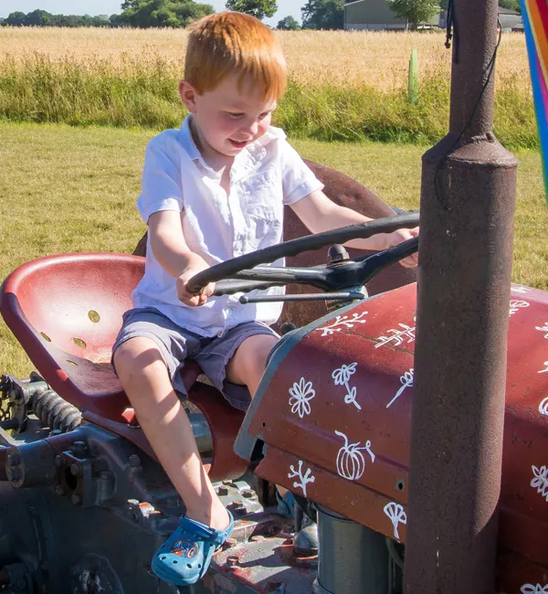 Young child with red hair sat on a toy tractor in a field, holding the steering wheel.